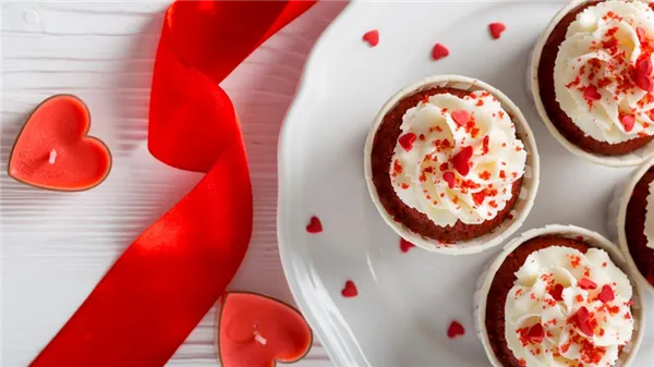 Капкейки «Красный бархат» на тарелке декорированные посыпкой в виде сердечек. Рядом свечи и красная лента